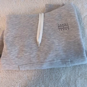 Grå hoodie från Tommy Jeans - En bekväm grå hoodie från Tommy Jeans med en vit dragsko och en känguruficka framtill. Hoodien har en tryckt logotyp på bröstet.