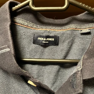 Grå t-shirt från Jack & Jones - En grå t-shirt från Jack & Jones med korta ärmar och en klassisk krage med knappar. T-shirten har en enkel design och är tillverkad i ett bekvämt material.