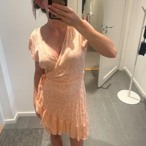 Ljus orange blommig klänning från BikBok - En ljus orange blommig klänning från BikBok i storlek S. Klänningen har korta ärmar, omlott-design och volangdetaljer vid fållen. Den är knälång och har en knytning i midjan.