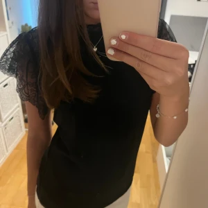Svart topp med spetsärmar - En elegant svart topp med spetsärmar. Toppen har en normal passform och är perfekt för festliga tillfällen eller i vardagen.