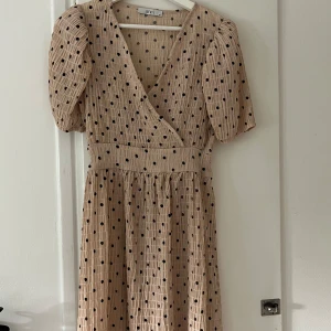 Beige prickig klänning - En beige klänning med svarta prickar. Klänningen har korta puffärmar och en omlott-design framtill. Den har en markerad midja och en kjol som faller löst.