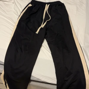 Svarta mjukisbyxor med vita ränder - Ett par svarta mjukisbyxor med vita ränder längs sidorna. Byxorna har en elastisk midja med dragsko för justering.