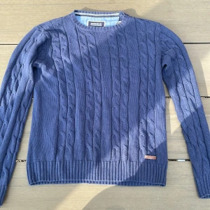 Blå stickad tröja från Bondelid - En blå stickad tröja från Bondelid med ett kabelstickat mönster. Tröjan har långa ärmar och ribbstickade muddar vid ärmslut och nederkant.