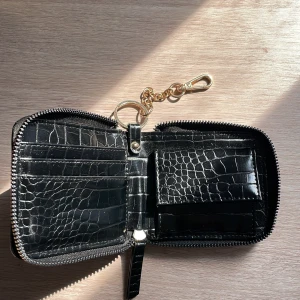 Svart plånbok med krokodilpräglat mönster - En elegant svart plånbok med krokodilpräglat mönster. Plånboken har en dragkedja runtom och en guldfärgad kedja med karbinhake. Insidan har flera fack för kort och en knappstängd ficka för mynt.