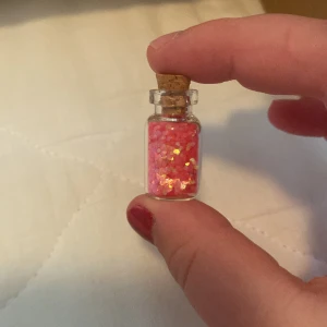 Rosa glitter i glasflaska - En liten glasflaska med kork som innehåller rosa glitter. Glittret har en skimrande effekt med inslag av guld och silver.