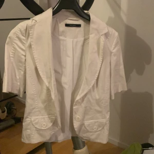 Vit kavaj från Vero Moda - En stilren vit kavaj från Vero Moda med korta ärmar och två knappar framtill. Kavajen har två fickor med lock och är tillverkad i ett lätt material, perfekt för vår och sommar.