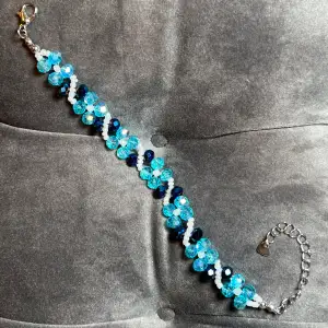 Glittriga blåa och vita pärlor i fint mönster med ett silvrigt spänne. Justerbar passform mellan 18-24 cm.