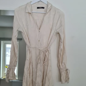 Gräddvit satinklänning  - Gräddvit satinklänning, knyts i midjan och runt handlederna. Behöver absolut strykas/steamas. Aldrig använd, nypris 499:-, normal i storleken. Kom med prisförslag 😊 Har även en likadan i brunt till försäljning.  Material: 100% Viskos. 