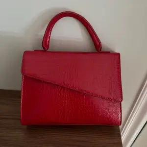 En röd handväska i skinn. Medföljer ett justerbart axelband. 