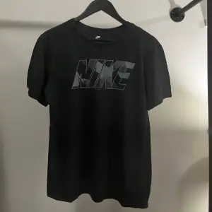 En svart t-shirt från Nike med ett stort Nike-logotyptryck i grått på framsidan. T-shirten har korta ärmar och en normal passform. Tillverkad i bomull.