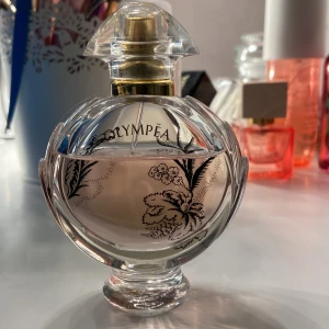 Olympea parfym - En elegant parfymflaska från Olympea med en rund form och genomskinligt glas. Flaskan har ett vackert blommönster och en gyllene spraypump. Parfymen inuti är ljusrosa. Ca 80% kvar i