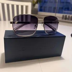 Ett par eleganta solglasögon från Louis Vuitton med en rektangulär form. Glasögonen har en silverfärgad metallram och lila tonade linser. Skalmarna har ett diskret rutmönster och Louis Vuitton-logotypen är synlig på linsen och skalmen.