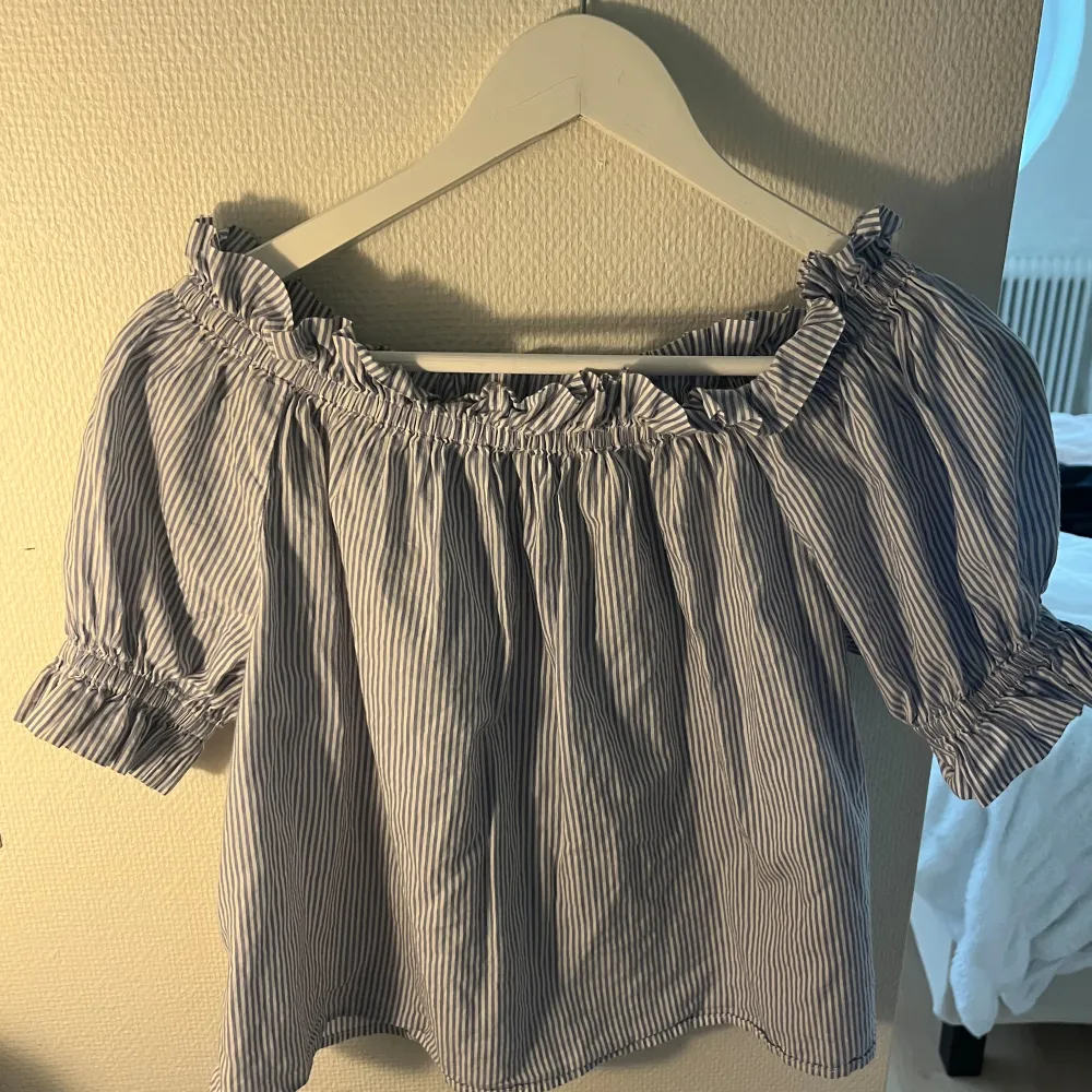 En blå- och vitrandig offshoulder-top med korta ärmar. Toppen har elastiska band runt axlarna och ärmsluten, vilket ger en rynkad effekt.. Toppar.