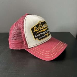 Röd och vit keps från Stetson - En röd och vit keps från Stetson med ett nätmaterial på baksidan och ett broderat märke på framsidan. Kepsen har en böjd skärm och justerbar passform. Skicket är 10/10 eftersom jag bara användt den nån enstaka gång!