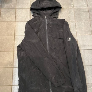 Legacy windbreaker. - Galet snygg windbreaker från märket legacy. Perfekt skick med en snygg logga på armen och band för att spänna åt luvan samt text på dragkedjan. Den är svart med en design så att den nästan ser lite camo ut med olika nyanser av svart. Nypris: 1200kr