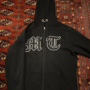 Svart hoodie med dragkedja och tryck - Säljer en svart hoodie med dragkedja och tryck på framsidan. Den har en stor huva med snören och långa ärmar. Perfekt för höst och vinter, den är både varm och bekväm. Passar bra till vardags eller chillkvällar.