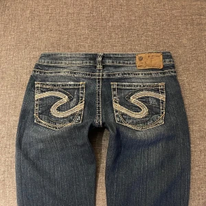 Lågmidjade bootcut jeans - Säljer ett par snygga blå jeans med broderade bakfickor och vita stygn. De har en låg midja och en bootcut-stil. Jeansen har slitna detaljer på framsidan och är i fint skick! Midjemått 38 Innerbenslängd 83. 💕 Jag är 165