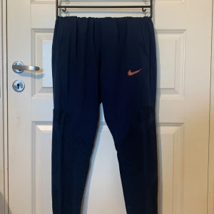 Mörkblå träningsbyxor från Nike - Säljer ett par mörkblå träningsbyxor från Nike i storlek L. Byxorna är slim fit och har en elastisk midja. De är tillverkade i Dri-FIT material som håller dig torr och bekväm under träning. Byxorna har en cool Nike-logga i rött och gult på ena benet. Perfekta för träning eller en avslappnad dag.