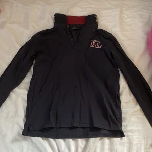 Svart långärmad tröja från Kingsland - Säljer en svart/marinblå långärmad rid-tröja från Kingsland Equestrian. Tröjan har en hög krage med knappar och ett broderat 'KL' märke på bröstet. Den är i bra skick och perfekt för ridning. Kragen har också 'Kingsland' broderat i vinrött och vitt. Materialet är bekvämt att ha på sig.