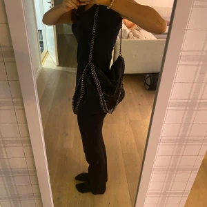 Svart axelväska med kedjedetaljer - Säljer en snygg svart axelväska med coola kedjedetaljer. Väskan är i syntetmaterial och har en glittrig yta som gör den perfekt för fest. Den har en kedja som axelrem och är rymlig nog för att få plats med allt du behöver. Perfekt för att ge din outfit en extra touch!