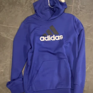Blå hoodie från Adidas - Säljer en snygg blå hoodie från Adidas. Den är i storlek XL och har en stor Adidas-logga på framsidan. Hoodien är perfekt för höst och vinter, och den är superbekväm med en stor ficka på magen. Den är i bra skick och redo för en ny ägare!