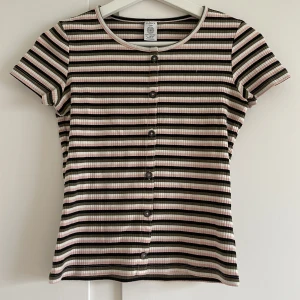 Randig ribbad topp från Lindex - Säljer en superfin randig topp från Lindex i storlek XS. Den är ribbad och har knappar hela vägen fram. Färgerna är en mix av rosa, svart, beige och vit. Perfekt för både vardag och fest! Toppen är i bra skick och väldigt bekväm att ha på sig.