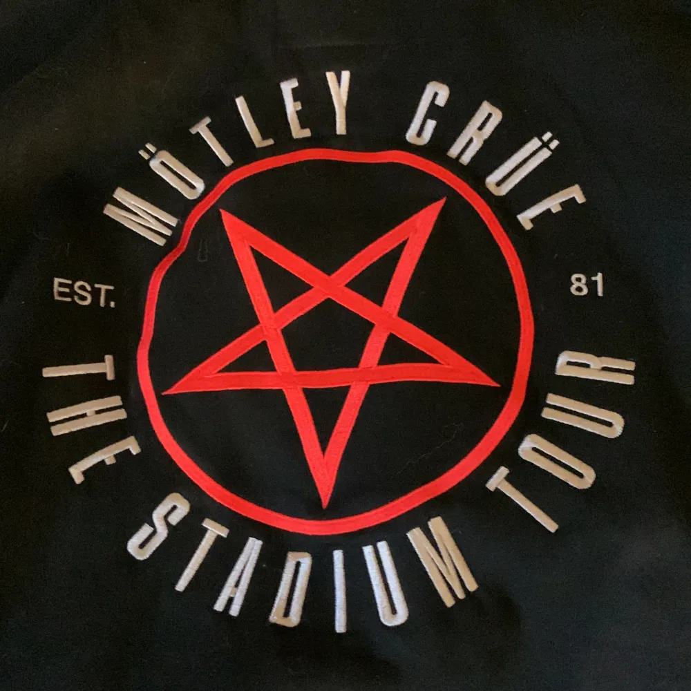 En svart jacka med huva från Mötley Crüe. Jackan har en dragkedja framtill och är dekorerad med bandets logotyp och texten 'The Stadium Tour'. Insidan av huvan är röd med en vit pentagramdesign. Jackan har långa ärmar och en ficka på vardera sida.. Jackor.