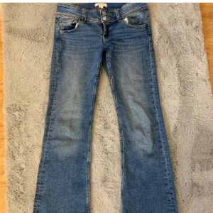 Blå jeans från Gina Tricot - Säljer ett par snygga blå jeans från Gina Tricot i storlek 152. De är lågmidjade och har en bootcut-stil. Jeansen är i bra skick och perfekta för både vardag och fest. De har knappar och dragkedja framtill samt fickor både fram och bak.