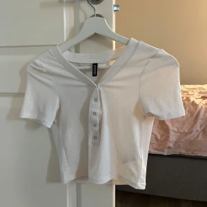 Vit ribbad topp från Gina Tricot - Säljer en vit ribbad topp från H&M. Den har en v-ringad hals och knappar framtill. Toppen är kortärmad och croppad. Använd ett fåtal gånger, så den är i bra skick. Perfekt för både vardag och fest!🤍