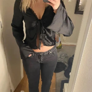 Svart knytblus med vida ärmar - Säljer en snygg svart blus med knytning framtill och vida ärmar. Blusen är croppad och har en fin spetsdetalj vid kanten. Perfekt för fest eller en utekväll! 🖤
