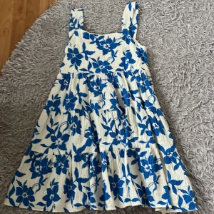 Blommig klänning från Zara - En vacker klänning från Zara med ett blommigt mönster i blått och vitt. Klänningen har breda axelband och är ärmlös. Den är tillverkad i ett lätt och bekvämt material, perfekt för varma sommardagar och är använd cirka 2 gånger!