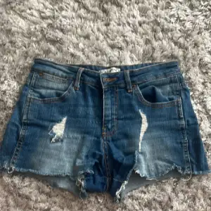 Säljer ett par snygga blå jeansshorts med slitningar fram och råa kanter. De har en klassisk femficksdesign och är perfekta för sommaren. Shortsen är i bra skick och passar perfekt för en avslappnad look.