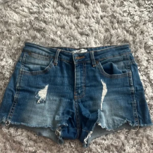 Blå jeansshorts med slitningar - Säljer ett par snygga blå jeansshorts med slitningar fram och råa kanter. De har en klassisk femficksdesign och är perfekta för sommaren. Shortsen är i bra skick och passar perfekt för en avslappnad look.