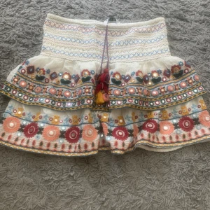 Vit broderad kjol med volanger - En vit kortkjol med broderade mönster i flera färger. Kjolen har volanger och dekorativa band med speglar och broderier. Den har även en knytning med tofsar i midjan. Från zara, säljs ej längre