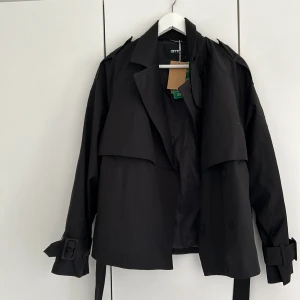 Svart kort trenchcoat  - Säljer denna oanvända korta trenchcoaten från aimn. Den har ett skärp i midjan men det går också att ta bort om man så vill det. Helt oanvänd med lapparna kvar! 🫶🏼 Orignalpriset låg mellan 699 och 999 kr