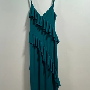 Blå/Grön långklänning - Långklänning i petroliumblå/grön med volanger och slits på höger sida. Klänningen har en insydd kort innerklänning. Jag är 164 cm och för mig passar den perfekt! Eftersom innerklänningen är kort bör den passa dig som är ca 160cm lång.