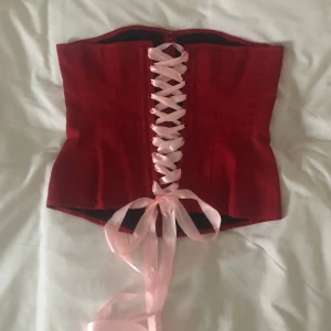 Röd korsett strl M/42 - Vintage korsett med boning i eldröd färg med rosa siden snörning. Passar större byst också. Midja som nu snörat: 86cm. Längd: 40cm Midja som snävast: 74cm. Passar större storlekar vid justering av snöre. Finns inga bilder på. Köparen står för frakt.