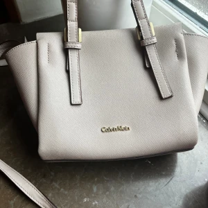 Calvin Klein väska - Söt handväska som kan användas över axeln/kroppen eller endast med de små handtagen. Det långa bandet går att ta av.   Använd men med försiktighet.  Se mått på sista bilden.  Rökfritt hem, hund finns 