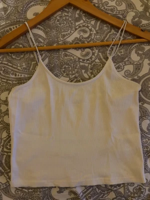 Vit ribbad linnetopp med smala band - Säljer en enkel och stilren vit ribbad linnetopp med tunna axelband. Toppen har en croppad passform och är perfekt för varma dagar eller som layering-piece. Materialet känns mjukt och stretchigt för extra komfort. Toppen är i storlek M/L.