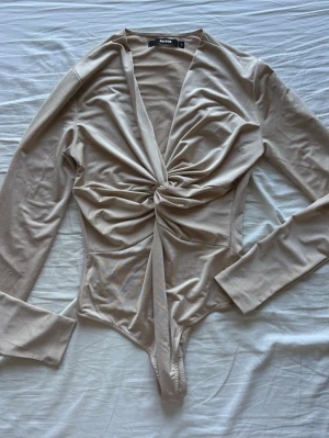 Beige body från Bikbok - Trendig beige body från Bikbok med lång ärm och twistad detalj framtill. Djup v-ringning och mjukt, stretchigt material 