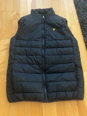 Svart dunväst från Lyle & Scott - Svart dunväst från Lyle & Scott med gul logga på bröstet. Västen har ståkrage, dragkedja framtill och horisontella sömmar som ger en snygg, puffig look. Perfekt för lager-på-lager och enkel att matcha med olika outfits.