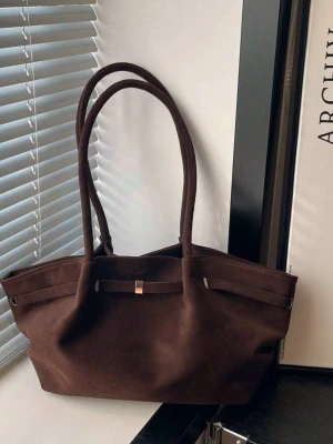 Mörkbrun mocka handväska - Snygg mörkbrun handväska i mjuk mocka med dubbla långa handtag och minimalistisk design. Väskan har en enkel gulddetalj framtill och rymlig insida, perfekt för dig som gillar stilrena accessoarer.