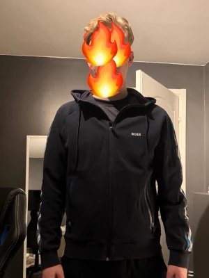 Mörkblå hoodie från BOSS med dragkedja - Snygg  hoodie från BOSS med dragkedja framtill och huva. Diskret BOSS-logga på bröstet och coolt mönstrad blå/vit rand längs ärmarna. Två fickor med dragkedja och ribbade muddar. Perfekt för en chill och stilren look.