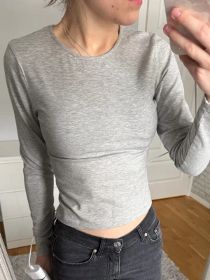 Ljusgrå croppad topp från Nelly - Säljer en ljusgrå croppad långärmad topp från Nelly. Toppen har en enkel och stilren design med rund halsringning och tight passform. Perfekt att matcha med jeans eller kjol för en clean look.