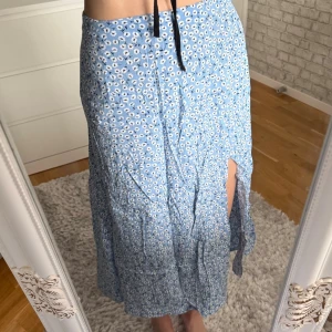 Blå omlottkjol med blommigt mönster - Snygg blå omlottkjol med vitt blommigt mönster och knytband i midjan. Kjolen har en slits på sidan som ger en trendig look. Perfekt för dig som gillar en feminin och avslappnad stil. Observera att den ej är struken på bilden!