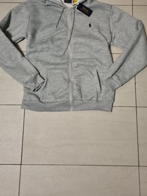 Grå zip hoodie från Polo Ralph Lauren - Snygg ljusgrå hoodie med dragkedja från Polo Ralph Lauren. Klassisk design med huva och två fickor framtill. Liten svart logga broderad på bröstet. Tillverkad i mjukt bomullsmaterial som känns skönt mot huden. Perfekt för en avslappnad och stilren look. Den känns som M! 