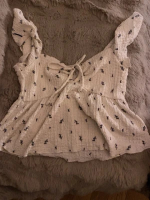 Vit blommig blus med snörning - Supersöt vit blus med små blå blommor och snörning framtill. Blusen har volangiga axelband och är tillverkad i ett krinklat bomullstyg som känns luftigt och somrigt. Perfekt för dig som gillar romantiska detaljer och en feminin stil.