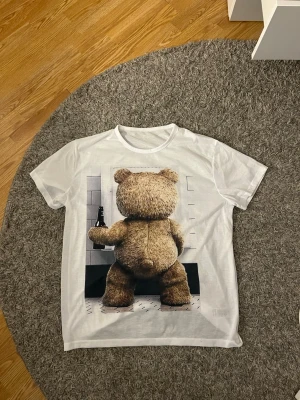 Vit t-shirt med coolt teddy Björn tryck. - Jag säljer en oversized t-shirt i väldigt bekvämt o mjukt material. Motivet är en teddybjörn på toan med en flaska i handen. Tyvärr vet jag inte vart den är ifrån. Den är i väldigt bra skick då den knappt är använd. Kan passa både herr och dam. Den är i storlek XL//L. Säljes för 40kr, pris kan diskuteras💕