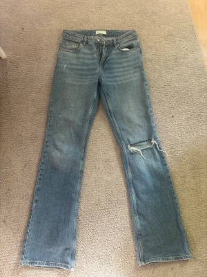 Blå raka jeans med slitning - Säljer ett par blå jeans med rak passform och cool slitning på ena knät. Jeansen har klassisk femficksdesign och en ljus tvätt som ger en avslappnad vibe. Perfekta för dig som gillar en trendig och avslappnad stil.