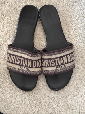 Christian Dior Paris sandaler - Snygga sandaler från Christian Dior med svart sula och breda remmar i beige och brunt tyg. Remmarna har tydlig logga med texten 'CHRISTIAN DIOR PARIS'. Perfekta för en trendig och avslappnad look.